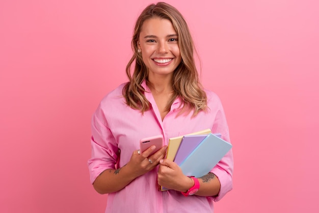 Blonde hübsche Frau in rosafarbenem Hemd lächelt, hält Notizbücher in der Hand und benutzt Smartphone