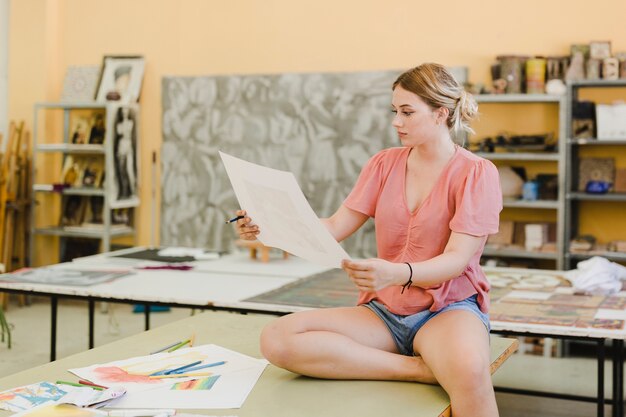 Blonde junge Frau, die auf dem Werktisch betrachtet ihre Malerei in der Werkstatt sitzt