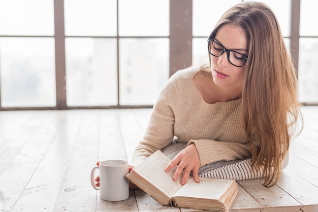 Blonde junge frau, die auf massivholzbodenlesebuch liegt