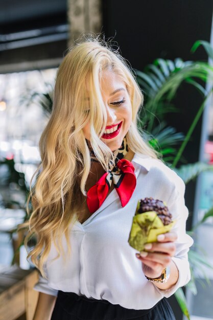 Blonde junge Frau, die gebackenes Schokoladenmuffin in der Hand betrachtet