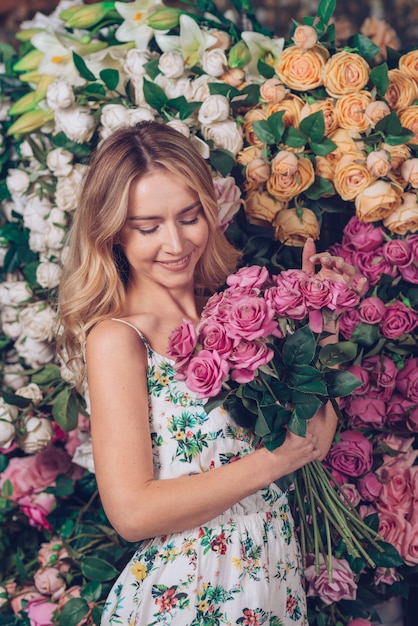Blonde junge Frau, die in der Hand rosa Rosen stehen vor Blumenhintergrund hält
