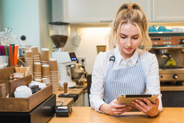 Blonde junge Frau, die in der Kaffeestube Theke betrachtet digitale Tablette steht