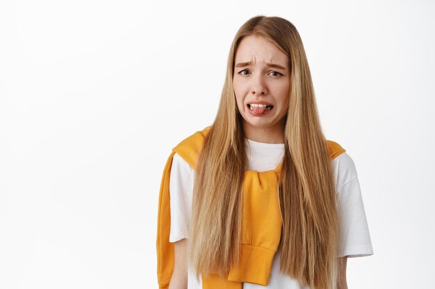 Blonde junge Frau, die sich angewidert fühlt, Zunge zeigt und aus Abneigung eine Grimasse verzieht, auf etwas ekelhaftes starrt, vor weißem Hintergrund steht