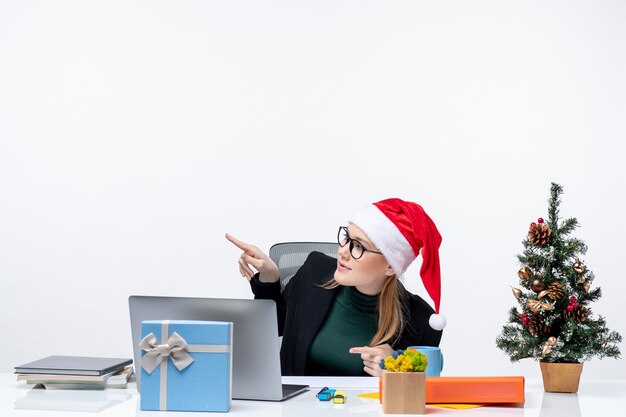 Blonde junge Frau mit Weihnachtsmannhut, der an einem Tisch mit einem Weihnachtsbaum und einem Geschenk darauf sitzt und oben auf weißem Hintergrund zeigt