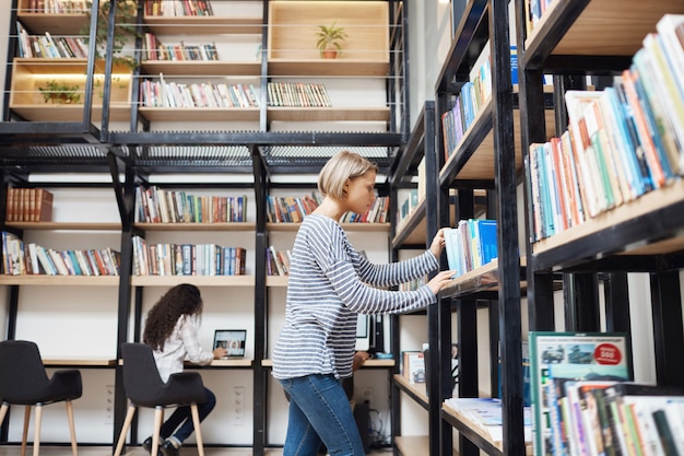 Kostenloses Foto blonde junge gutaussehende frau in gestreiftem hemd und jeans, die nach einem buch im regal in der bibliothek sucht und sich auf prüfungen in der universität vorbereitet