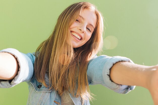 Blonde lächelnde junge Frau im Sonnenlicht, die selfie gegen grünen Hintergrund nimmt