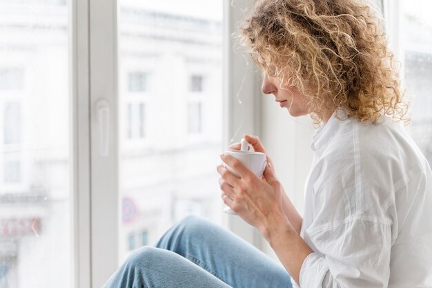 Blonde lockige Frau, die zu Hause in der Nähe des Fensters entspannt