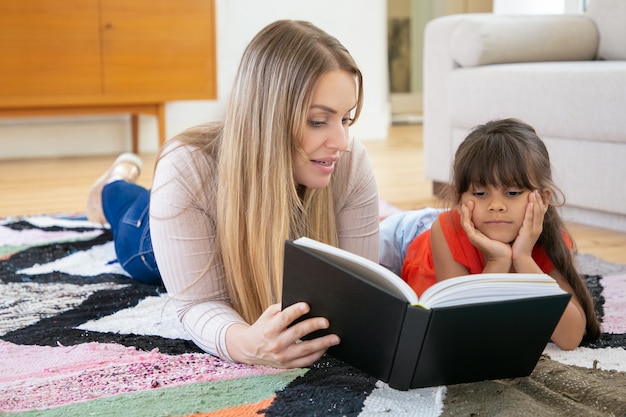 Blonde Mutter liegt auf Teppich mit Tochter und liest ihr ein Buch vor.