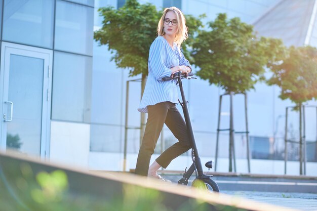 Blonde, nachdenkliche Frau mit Brille fährt ihren neuen Elektroroller an einem hellen, sonnigen Tag auf der Straße.