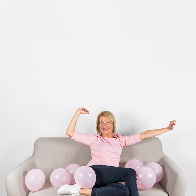 Blonde reife Frau, die auf Sofa mit Ballonen gegen weißen Hintergrund sitzt