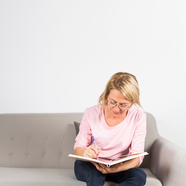 Blonde reife Frau, die auf Sofaschreiben im Tagebuch mit Stift sitzt