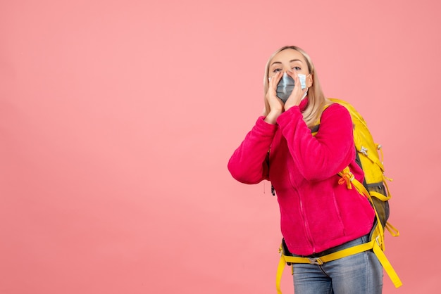 Blonde reisende Frau der Vorderansicht mit gelbem Rucksack, der Maske trägt, die jemanden anruft