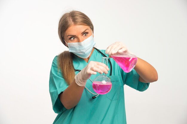 Blonde Ärztin, die Gesichtsmaske trägt und Chemikalien von einer Flasche zur anderen setzt.