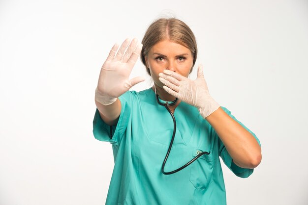 Blonde Ärztin in blauer Uniform mit Stethoskop im Nacken, das Schweigenzeichen macht.