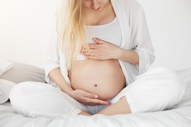 Blonde schwangere Frau, die ihren Bauch betrachtet und ihn mit ihren Händen stützt, die darauf warten, dass ihr Baby oder ihre Frau kommen.