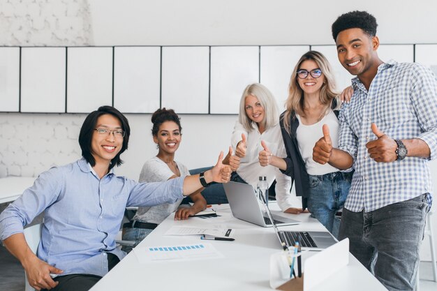 Blonde Sekretärin sitzt auf Tisch, während Büroangestellte mit Daumen hoch posieren. Innenporträt des glücklichen asiatischen Managers im trendigen Hemd, das im Konferenzsaal mit ausländischen Partnern lächelt.