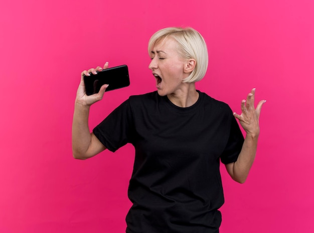 Blonde slawische Frau mittleren Alters, die mit geschlossenen Augen singt, die Hand in der Luft unter Verwendung des Mobiltelefons als Mikrofon lokalisiert auf rosa Wand halten