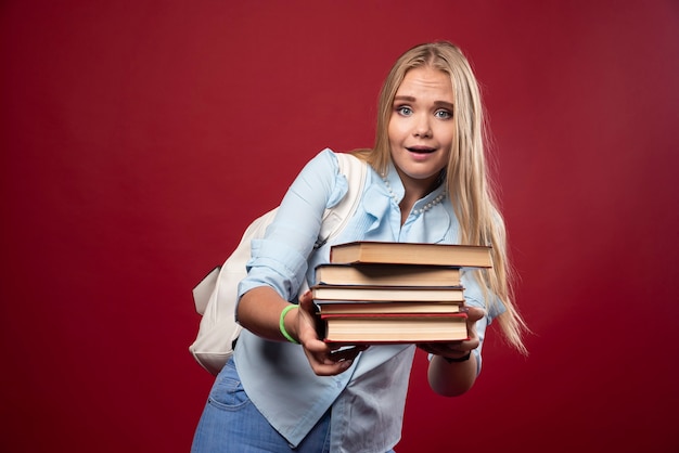 Blonde Studentin, die einen schweren Stapel Bücher hält und müde aussieht.