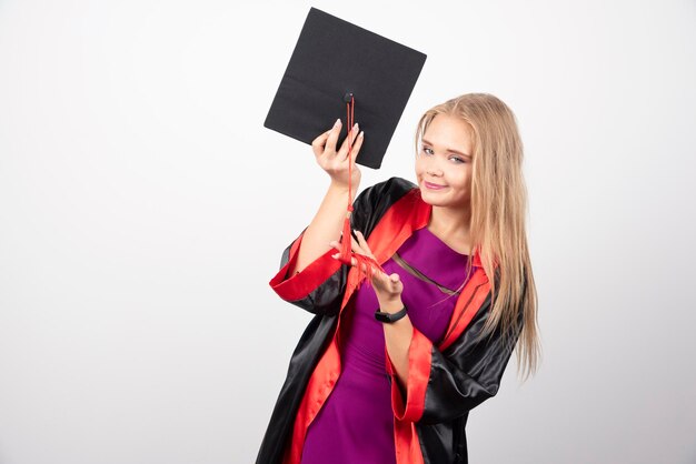 Blonde Studentin, die mit ihrer Kappe auf weißem Hintergrund aufwirft. Hochwertiges Foto