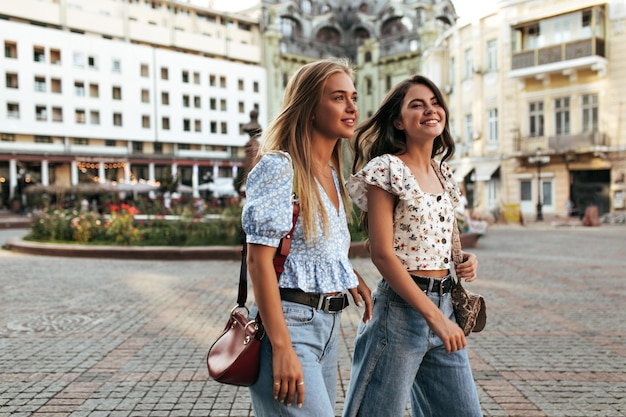 Blonde und brünette frauen in stylischen lockeren jeans und floralen, trendigen blusen gehen und unterhalten sich gut gelaunt draußen porträt attraktiver freundinnen