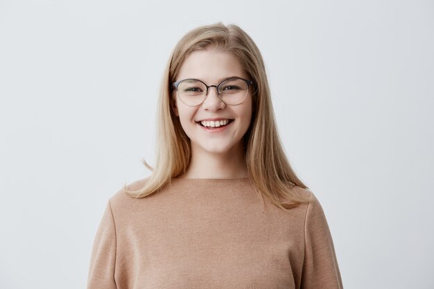 Blonde verspielte junge europäische Frau, die braunen übergroßen Pullover trägt, der über guten Witz lacht und perfekte weiße Zähne demonstriert, ihr Aussehen und Ausdruck voller Freude und Glück. Menschen und Lebensstil