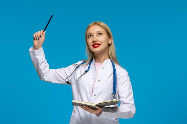 Blonder Arzt des Weltärztetages, der einen Stift und ein Notizbuch mit dem Stethoskop im medizinischen Kittel hält
