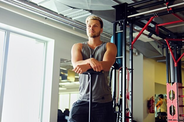 Blonder athletischer Mann in Sportbekleidung hält Langhantel über TRX-Standhintergrund in einem Fitnessclub.