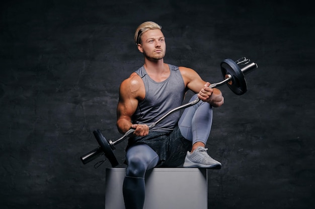 Blonder athletischer Mann in Sportbekleidung sitzt auf einer weißen Kiste und hält Langhantel.