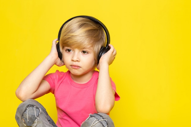 Blonder Junge in rosa T-Shirt und graue Jeans in schwarzen Kopfhörern, die Musik hören