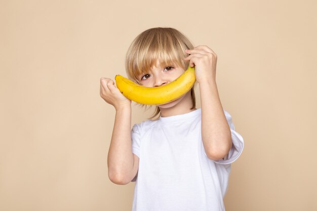 Blonder Junge lächelt süß süß mit Banane im weißen T-Shirt auf rosa Schreibtisch