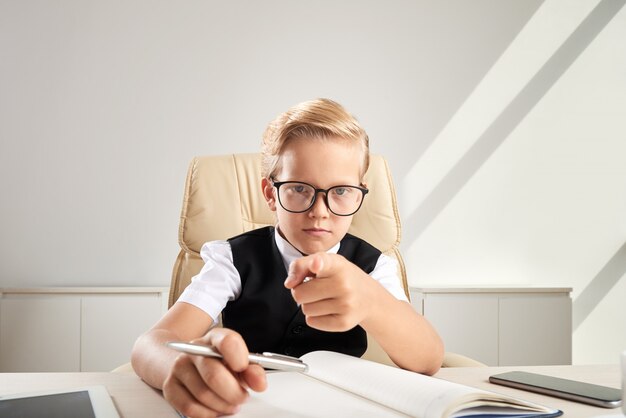 Blonder kaukasischer Junge in den Gläsern, die am Schreibtisch im Büro sitzen und in Richtung zur Kamera zeigen