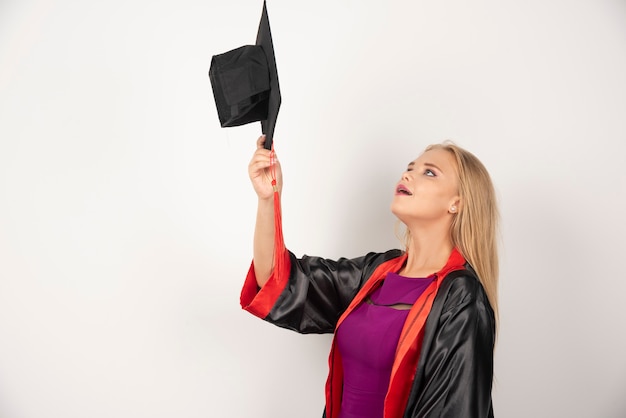 Blonder Student, der ihre Kappe auf Weiß betrachtet.