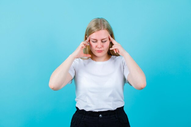 Blondes junges Mädchen denkt vorbei, indem es die Finger an den Schläfen auf blauem Hintergrund hält