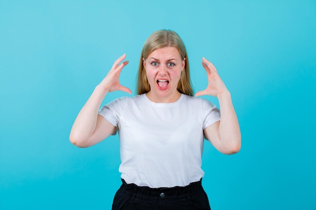 Blondes junges Mädchen schreit, indem es die Hände in der Nähe des Kopfes auf blauem Hintergrund hält