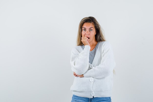 Blondes Mädchen beißt Fäuste, während es an etwas in gestreiftem T-Shirt, weißer Strickjacke und Jeanshose denkt und überrascht, Vorderansicht schaut.