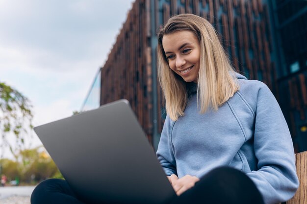 Blondes Mädchen, das auf Bank sitzt und Laptop hält