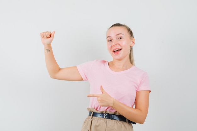 Blondes Mädchen, das auf etwas zeigt, das vorgab, im T-Shirt, in der Hose gehalten zu werden und glücklich zu sehen, Vorderansicht.