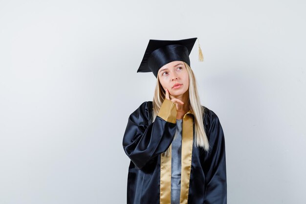 Blondes Mädchen, das aufschaut, während es in Absolventenuniform posiert und nachdenklich aussieht.