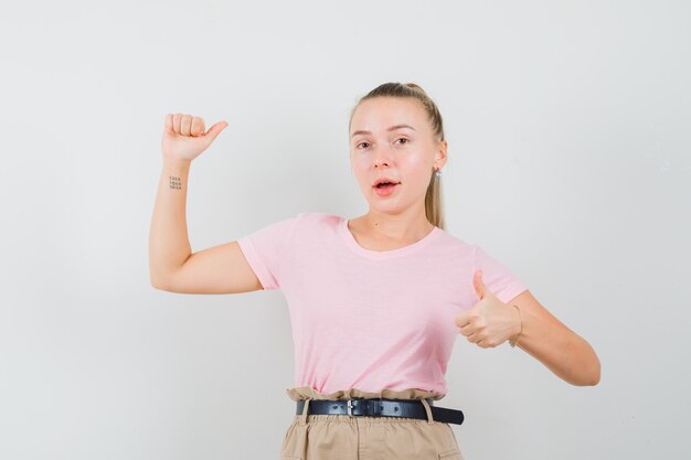 Blondes Mädchen, das Daumen oben zeigt, Arm im T-Shirt, in der Hose hebt und selbstbewusst aussieht. Vorderansicht.