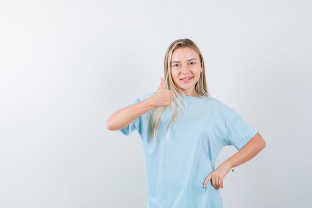 Blondes Mädchen, das Daumen oben zeigt, Hand auf Taille im blauen T-Shirt haltend und selbstbewusst, Vorderansicht schauend.