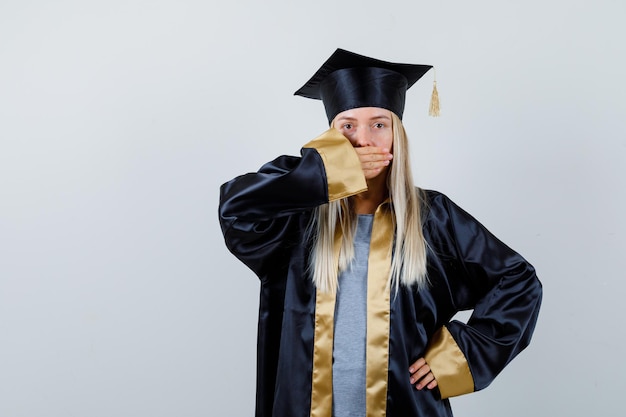 Kostenloses Foto blondes mädchen, das den mund mit der hand bedeckt, während es die hand an der taille in abschlusskleid und mütze hält und überrascht aussieht