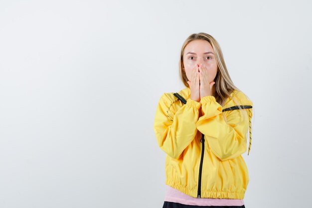 Blondes Mädchen, das die Hände in der gelben Jacke am Mund hält und verängstigt aussieht