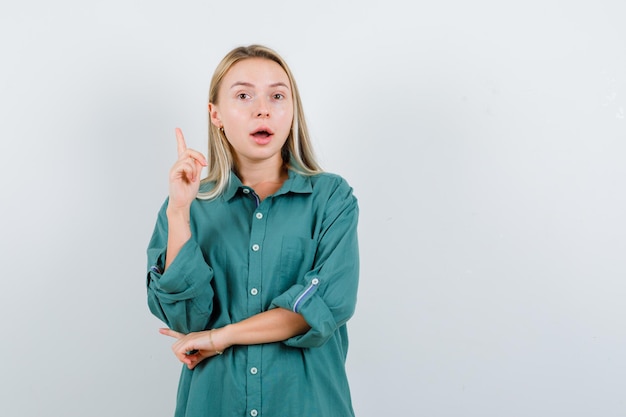 Blondes Mädchen, das eine Hand in Heureka-Geste hebt, während es die Hand am Ellbogen in grüner Bluse hält und vernünftig aussieht.