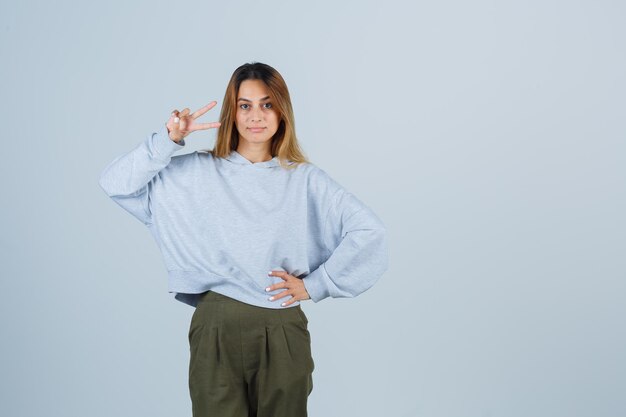 Blondes Mädchen, das Friedensgeste zeigt und Hand an der Taille in olivgrünem blauem Sweatshirt und Hosen hält und strahlend aussieht, Vorderansicht.