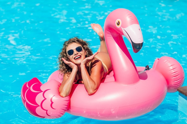 Kostenloses Foto blondes mädchen, das für instagram-geschichten posiert und sich im sommerpool auf einem aufblasbaren rosa flamingo in einem badeanzug ausruht.