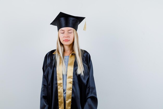 Blondes Mädchen, das gerade steht, die Augen schließt und in Abschlusskleid und Mütze vor der Kamera posiert und entspannt aussieht.