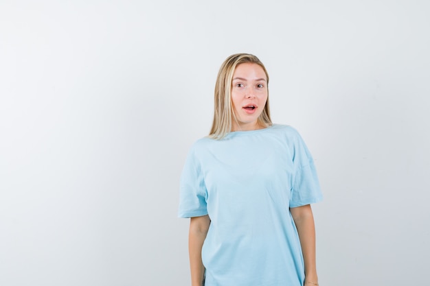 Kostenloses Foto blondes mädchen, das gerade steht und an der kamera im blauen t-shirt aufwirft und hübsch schaut, vorderansicht.