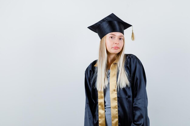 Blondes Mädchen, das gerade steht und in Abschlusskleid und Mütze vor der Kamera posiert und charmant aussieht