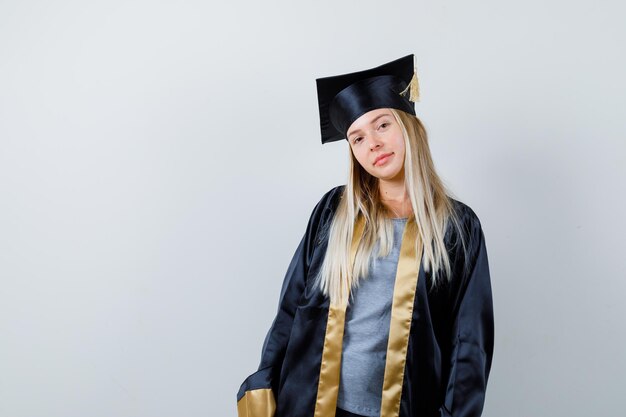 Blondes Mädchen, das gerade steht und in Abschlusskleid und Mütze vor der Kamera posiert und süß aussieht