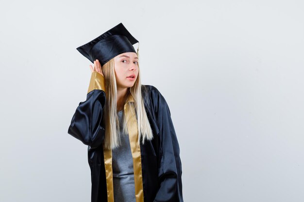 Blondes Mädchen, das Händchen in der Nähe des Ohrs hält, um etwas in Abschlusskleid und Mütze zu hören und fokussiert zu schauen.